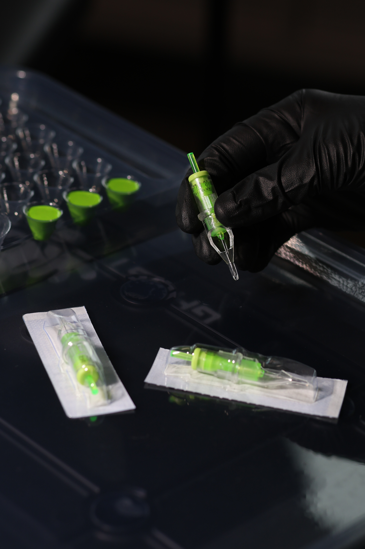 Gloved hand holding a green tattoo cartridge over a workstation
